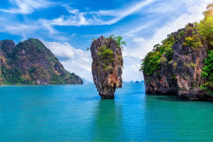 Tall rock formation in turquoise sea with lush islands under a blue sky.