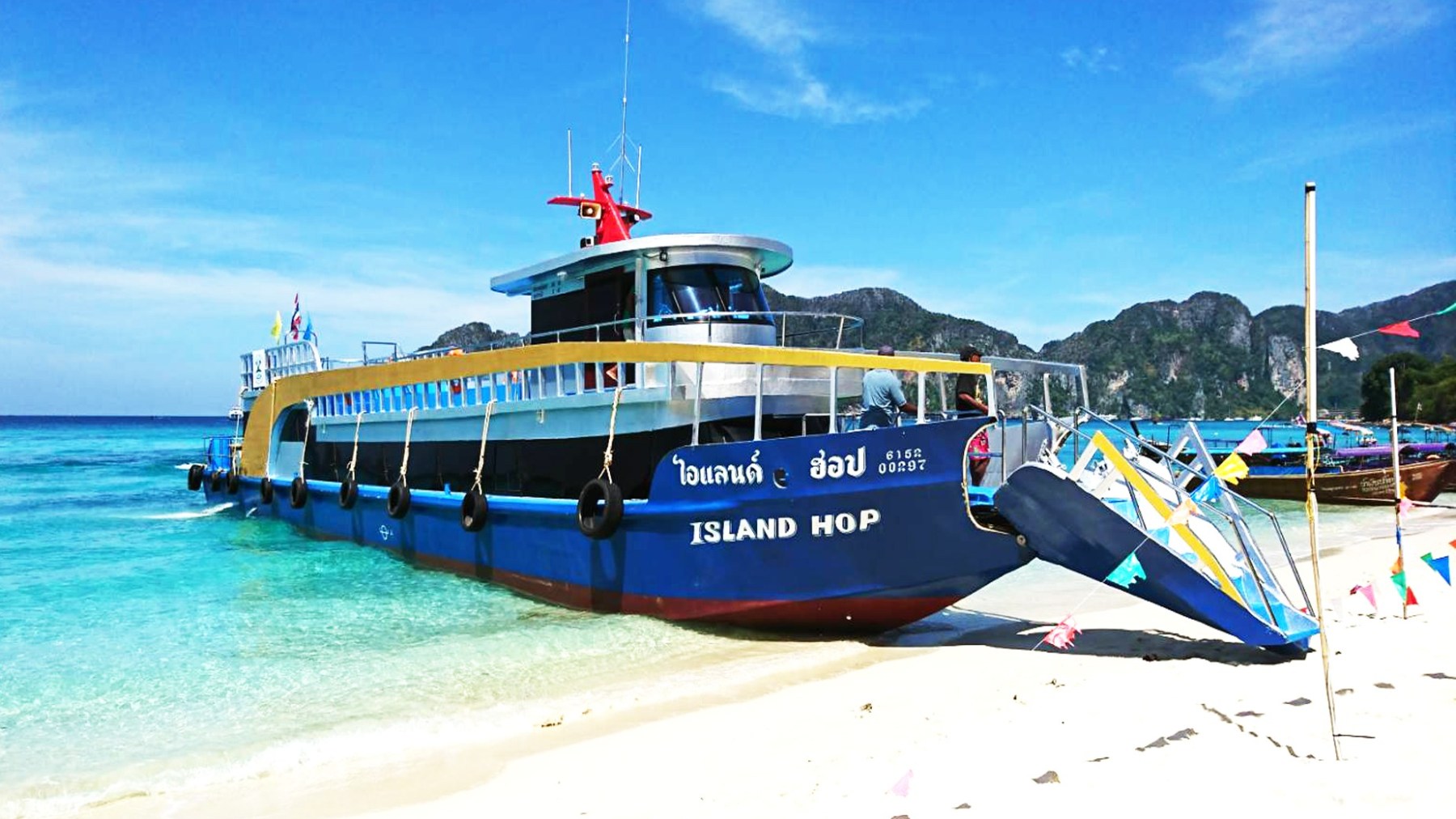 A blue ferry named 'Island Hop' docked on a tropical beach with mountains in the background.