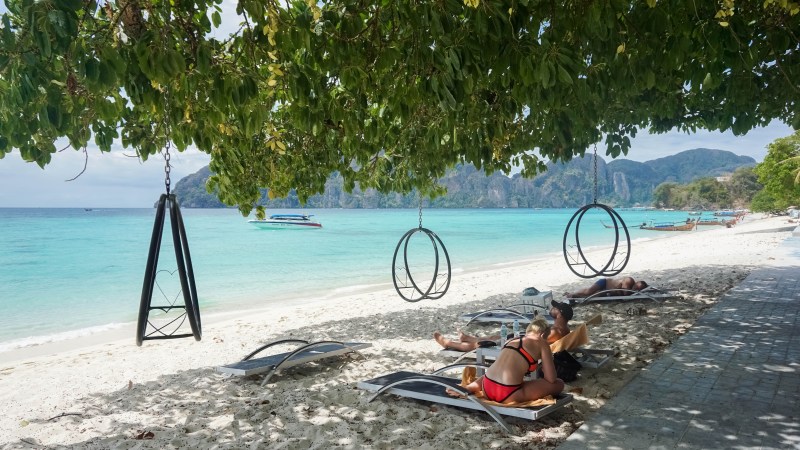 Beach scene with loungers, hanging rings, and boats on turquoise water under tree shade.