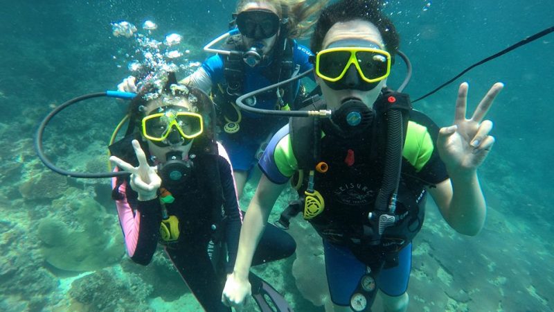 Three scuba divers underwater, wearing masks and gear, making peace signs.