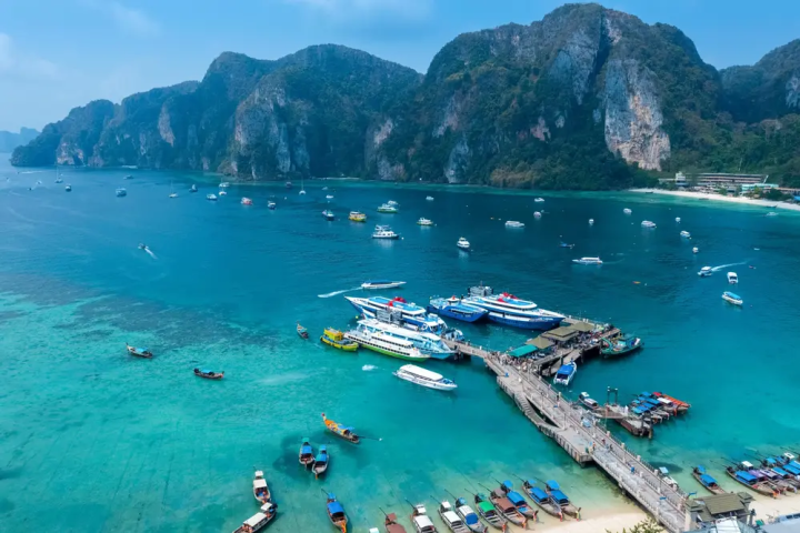 Aerial view of a turquoise bay with boats, a dock, and cliffs in the background.
