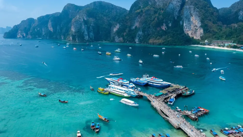 Aerial view of a turquoise bay with boats, a dock, and cliffs in the background.