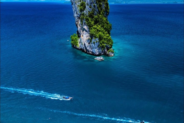 Lone rocky island with greenery in blue sea, two boats creating trails, clear sky in background.