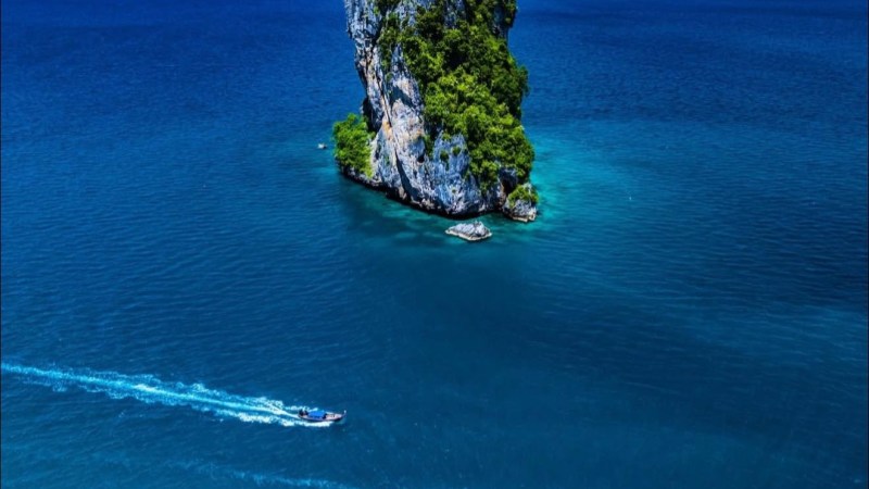 Lone rocky island with greenery in blue sea, two boats creating trails, clear sky in background.