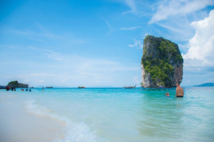 People swimming near a rocky island in clear blue water under a bright sky.
