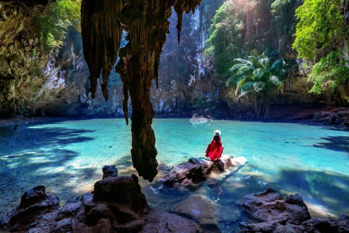 Person in red dress seated on rock by turquoise cave pool with sunlight streaming through.