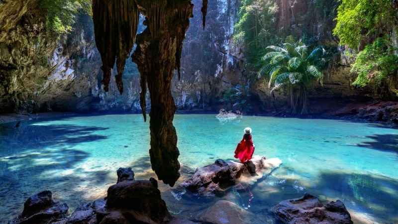 Person in red dress seated on rock by turquoise cave pool with sunlight streaming through.