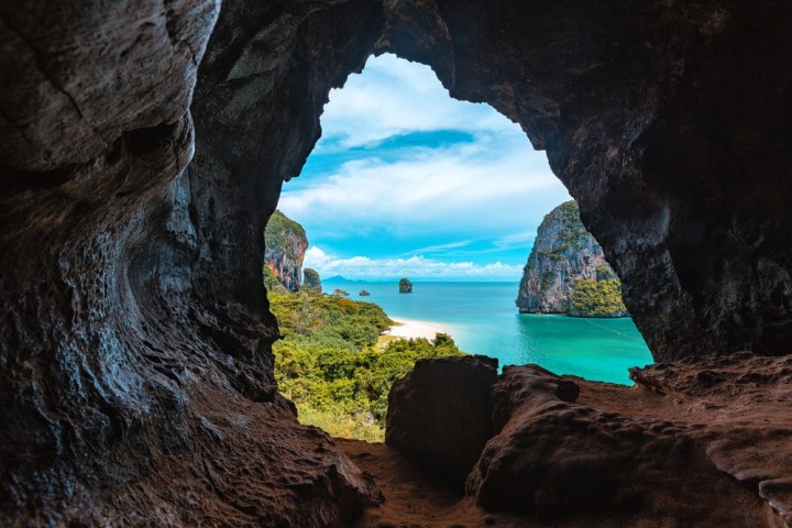 View through cave opening of turquoise sea and lush cliffs under a blue sky.