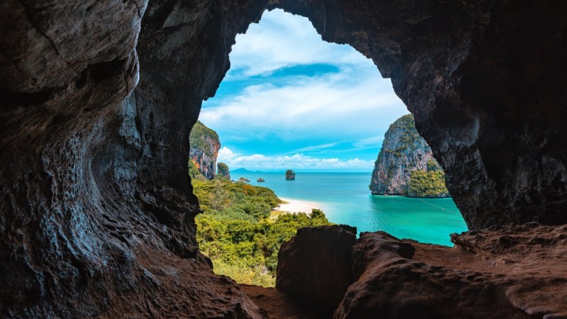 View through cave opening of turquoise sea and lush cliffs under a blue sky.