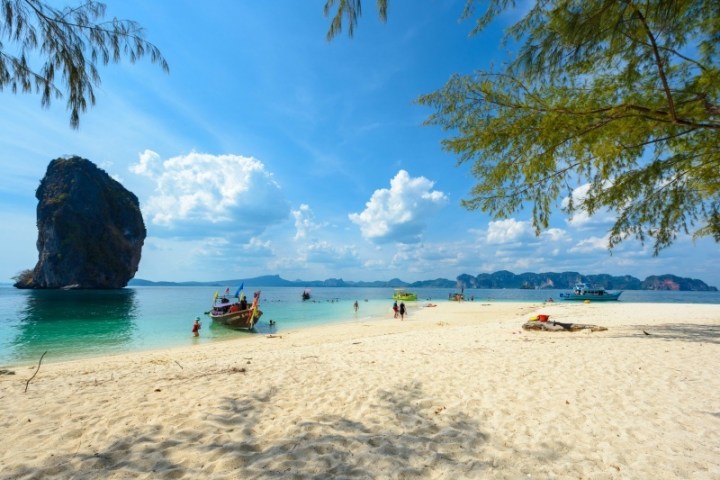 Beach with tourists, boats, and a tall rock formation under a blue sky with clouds.