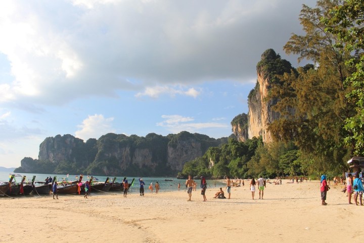 Sandy beach with people, longtail boats, and limestone cliffs under a cloudy sky.