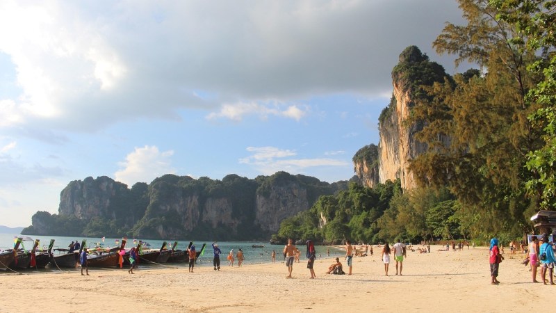 Sandy beach with people, longtail boats, and limestone cliffs under a cloudy sky.