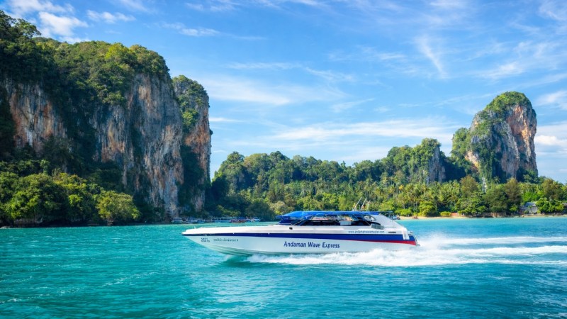A speedboat named Andaman Wave Express cruising on turquoise water with lush cliffs and blue sky in the background.