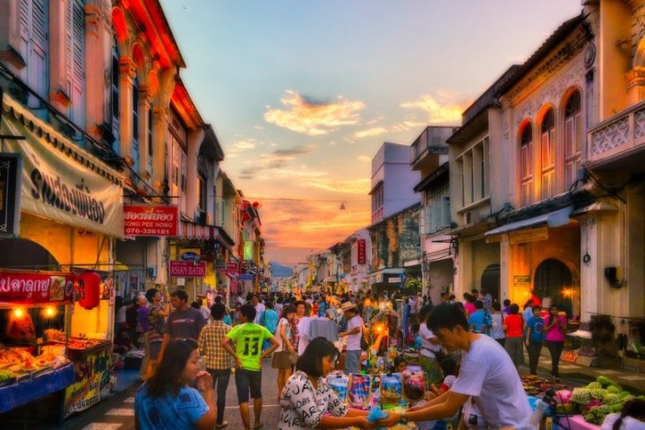 Bustling street market at sunset with people, colorful stalls, and vintage buildings.