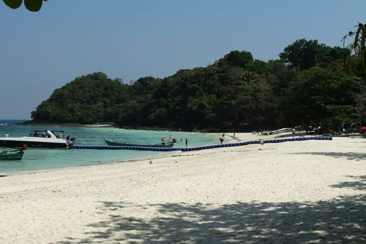 Tropical beach with boats, clear water, white sand, and lush green trees under a blue sky.