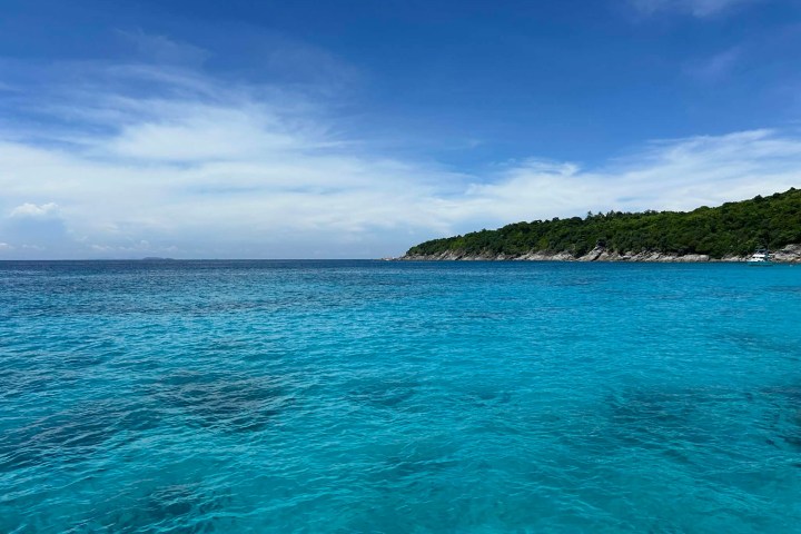 Clear blue ocean water with distant green shoreline under a blue sky with wispy clouds.