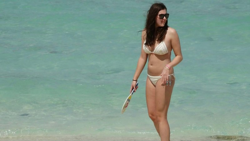 Woman in a white bikini holding a paddle, standing on a beach by the sea.