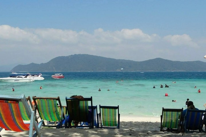 Beach scene with colorful chairs, people swimming, and boats on turquoise water under a clear sky.