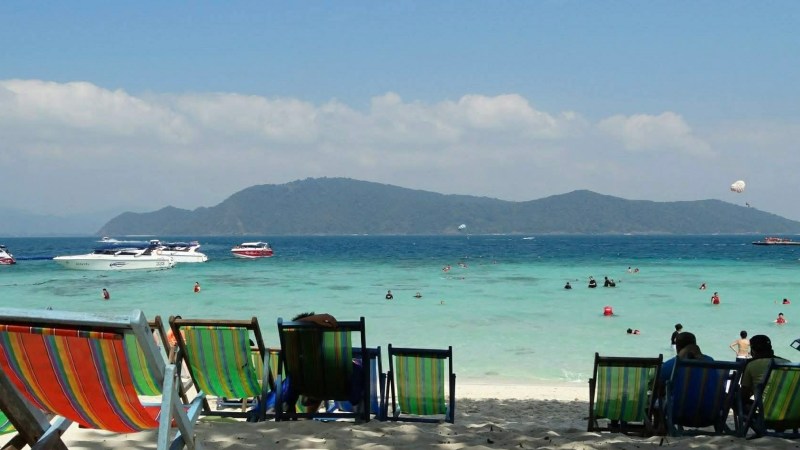 Beach scene with colorful chairs, people swimming, and boats on turquoise water under a clear sky.