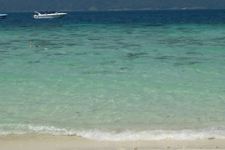 Turquoise sea with waves and boats in the distance near a sandy shore.