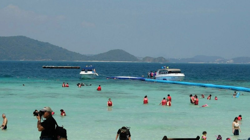 People swimming near boats in a tropical beach setting with distant hills.