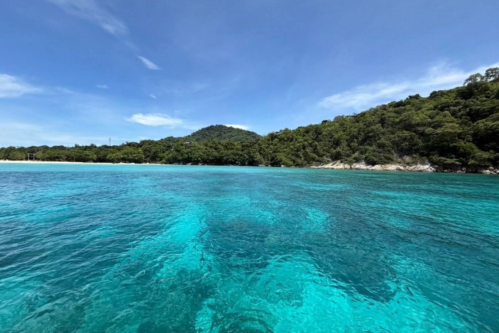 Clear turquoise water with lush green trees and a hill under a blue sky.