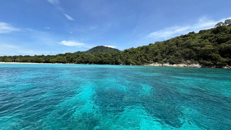 Clear turquoise water with lush green trees and a hill under a blue sky.