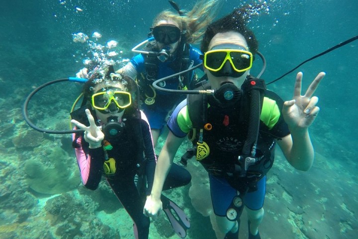 Three scuba divers underwater, making peace signs with coral in the background.