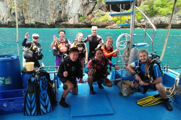 Group of scuba divers posing on a boat with rocky cliffs and ocean in the background.