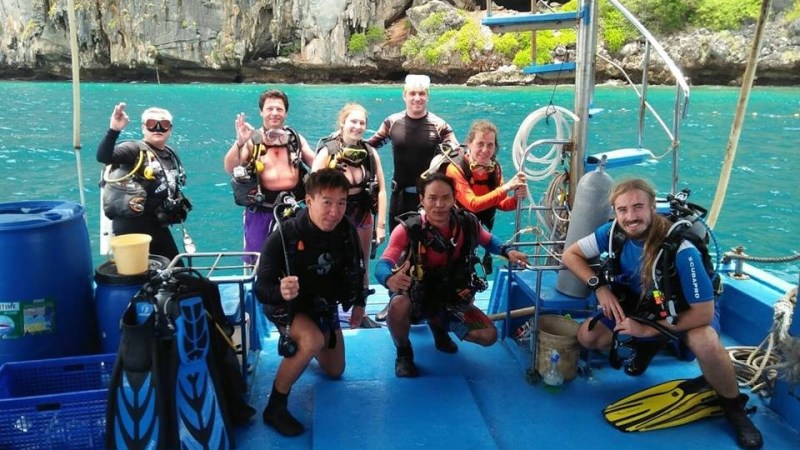 Group of scuba divers posing on a boat with rocky cliffs and ocean in the background.
