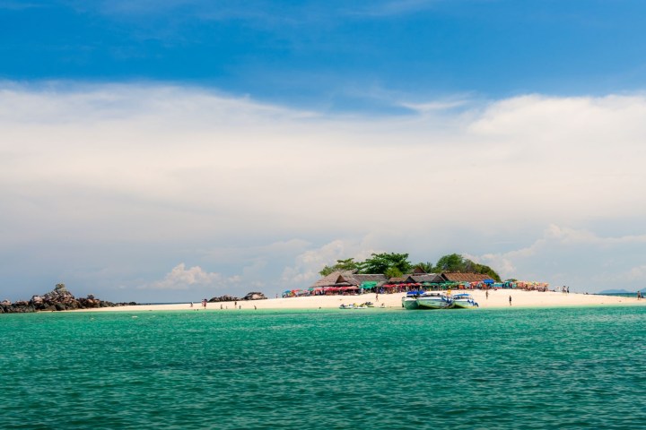 Tropical island with white sandy beach, boats, and clear turquoise water under a blue sky.