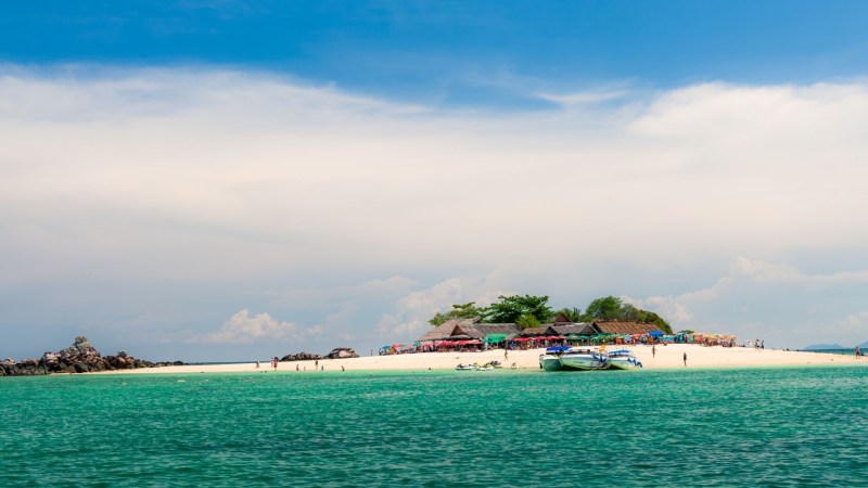 Tropical island with white sandy beach, boats, and clear turquoise water under a blue sky.