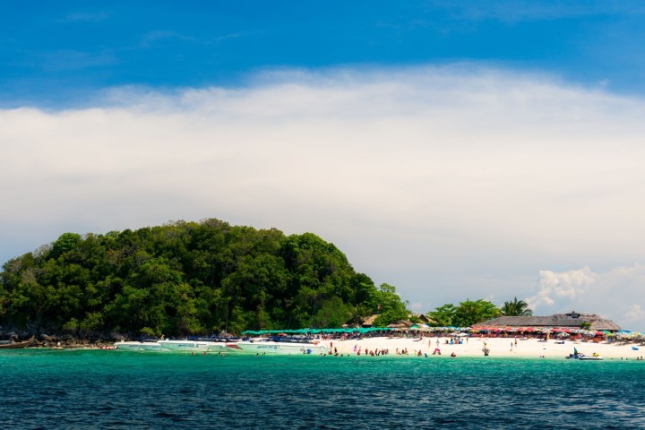 Tropical island with trees, white sandy beach, and people under colorful umbrellas, surrounded by blue sea.