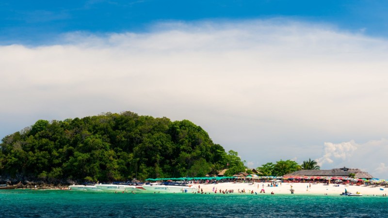 Tropical island with trees, white sandy beach, and people under colorful umbrellas, surrounded by blue sea.
