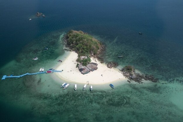 Aerial view of a small island with boats docked on clear water near sandy beach and lush vegetation.