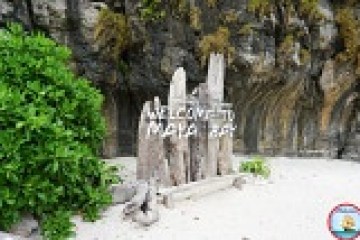 Driftwood sign reading 'Welcome to Maya Bay' on a sandy beach with rocky cliff and greenery.