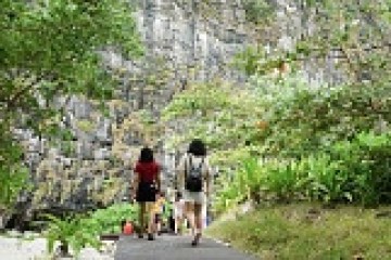 Two people walking on a path surrounded by greenery and a rocky cliff.