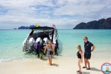 Tourists disembark a speedboat on a tropical beach with clear water and islands in the background.