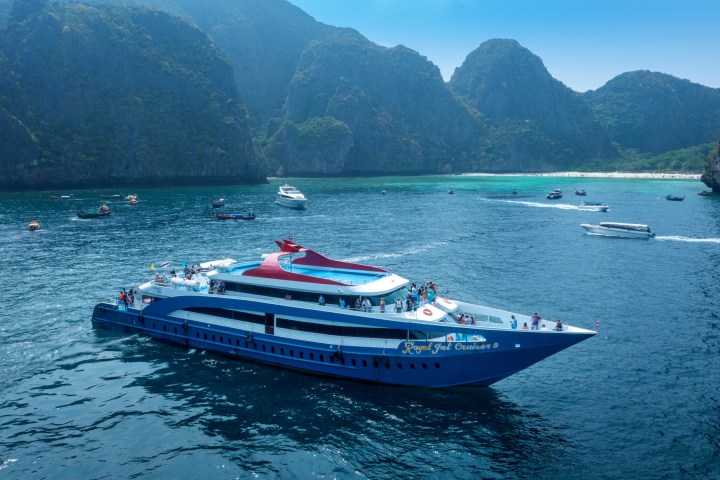 Large blue cruise ship in ocean with people onboard, surrounded by smaller boats and rocky islands in background.