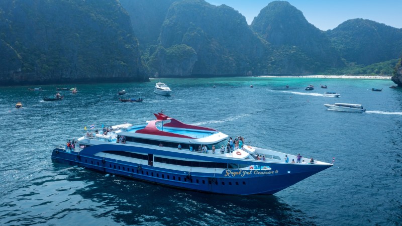 Large blue cruise ship in ocean with people onboard, surrounded by smaller boats and rocky islands in background.