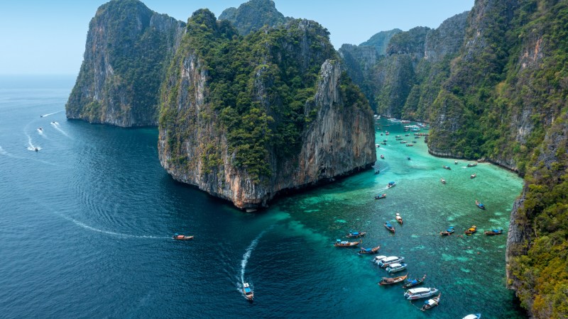 Aerial view of turquoise bay with boats and lush cliffs.