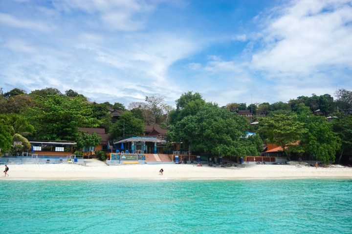 Scenic beach with turquoise water, trees, and buildings in the background.