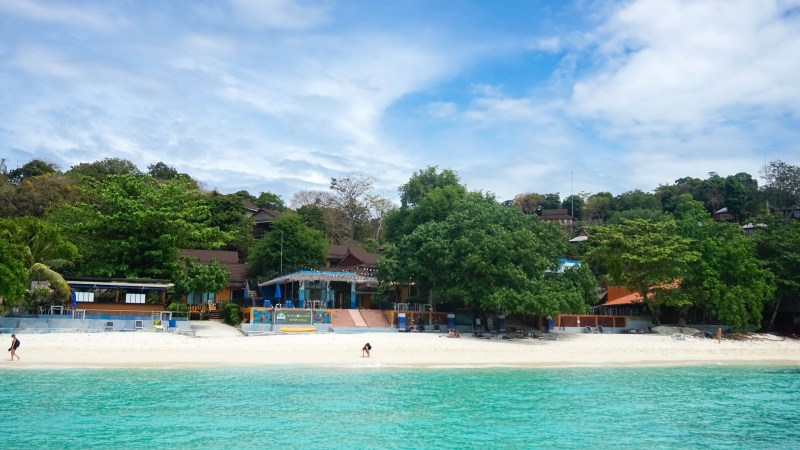 Scenic beach with turquoise water, trees, and buildings in the background.