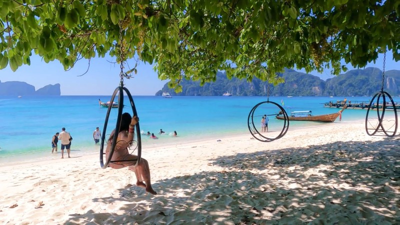 People enjoying beach with swings, turquoise sea, and boats.