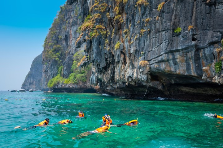 Snorkelers in orange vests swim near a steep, rocky cliff with clear turquoise water.