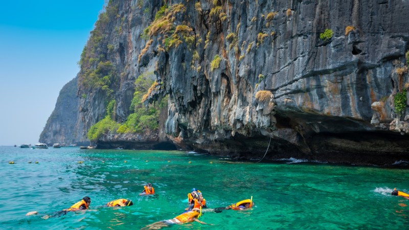 Snorkelers in orange vests swim near a steep, rocky cliff with clear turquoise water.