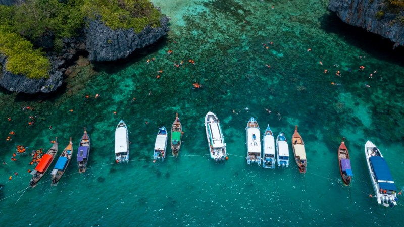 Aerial view of boats lined in turquoise water near rocky cliffs.