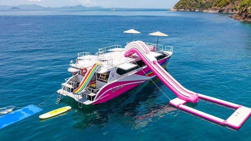 Colorful yacht with slides and inflatable platform floating in clear blue water near a rocky shore.