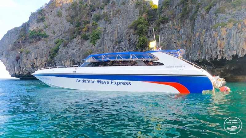 White and blue speedboat 'Andaman Wave Express' near a rocky cliff in clear turquoise water.