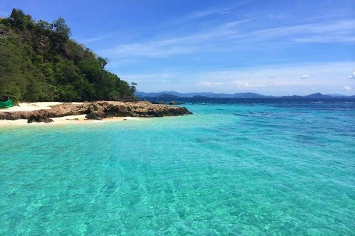 Tropical beach with clear turquoise water, rocky shoreline, and lush greenery under a blue sky.